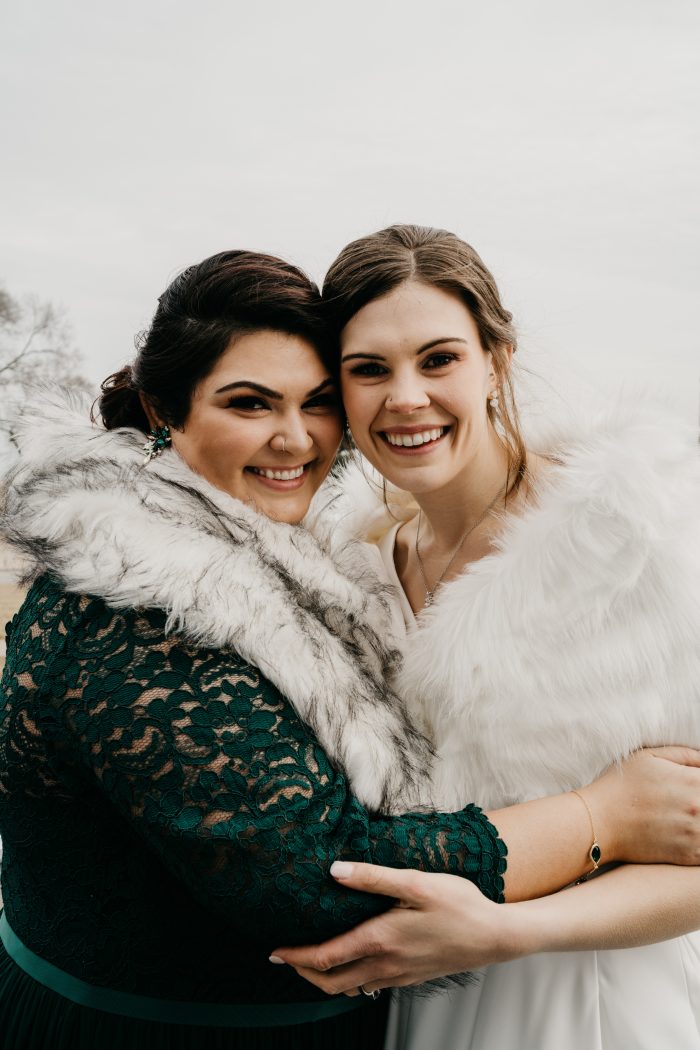 Bridesmaid in green lace dress with a fur wrap hugging around the bride wearing a fur wrap